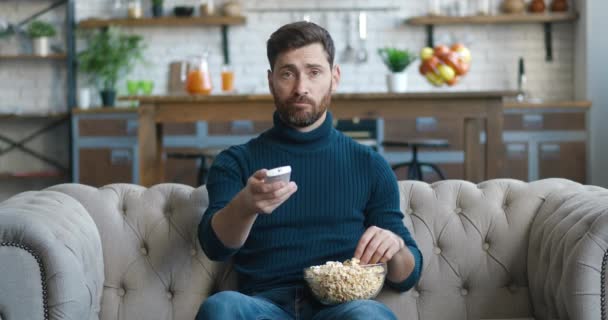 Junger bärtiger Mann sitzt zu Hause auf der Couch und schaut fern. Gelangweilte Millennialmännchen nutzt die Fernbedienung, um auf der Suche nach interessanten Videoinhalten die Kanäle zu wechseln und isst Popcorn