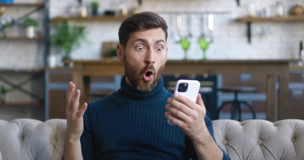 Emocionado y agradablemente sorprendido hombre guapo se regocija por el mensaje en su teléfono inteligente. Hombre joven mirando el teléfono móvil y se siente sorprendido feliz y diciendo WOW en el fondo del hogar. — Vídeo de stock