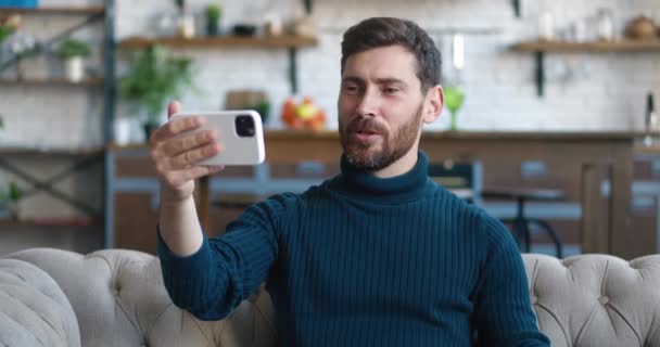 Jeune homme barbu beau tenant smartphone dans le bras tendu, faire une conférence par appel vidéo, sourire saluant son interlocuteur, assis sur le canapé à la maison. Concept de communication en ligne — Video