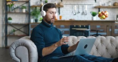 Kulaklıklı sakallı bir iş adamı, video görüşmeleri, evdeki kanepede oturuyor. Çevrimiçi eğitim, mesafe eğitimi, iş toplantısı, Webinar konferansı.