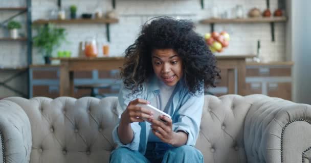 Фатальна афро-американська жінка, яка грає захоплюючий мобільний додаток на своєму смартфоні. Розчарована жінка, яка втратила гоночну гру, засмучена тим, що загубила гру, сидячи на дивані у вітальні вдома.. — стокове відео