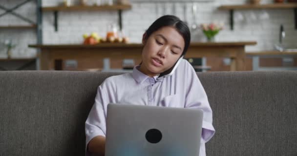 Busy asian business woman professional talking on phone using laptop sitting on sofa at living room, happy female freelancer working distantly from home. Communication and business people concept. — Stock Video