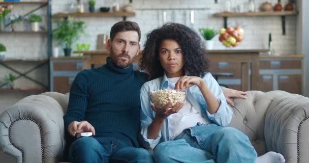 Joven familia interracial viendo la película de comedia en la televisión con palomitas de maíz sentado en el sofá en el apartamento. Riendo pareja viendo divertido vídeo juntos en la televisión en casa — Vídeos de Stock
