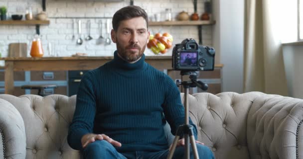 自宅でプロのカメラで新しいvlogビデオを撮影人気ブロガーの男.男性コンテンツ作成者はソファに座ってオンラインレッスンを撮影します。ビデオカメラでvlogを記録するVlogger — ストック動画