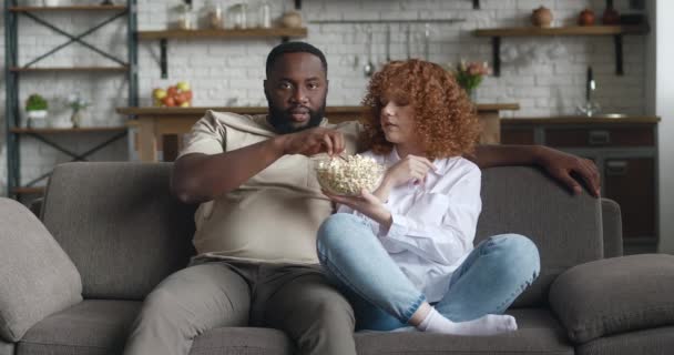 Joven familia interracial viendo la película de comedia en la televisión con palomitas de maíz sentado en el sofá en el apartamento. Riendo pareja viendo divertido vídeo juntos en la televisión en casa — Vídeos de Stock