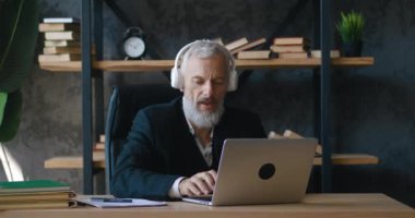 Ofisteki dizüstü bilgisayarda çalışırken kulaklıkla müzik dinleyen mutlu sakallı yetişkin iş adamı.