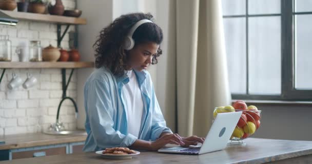 戴耳机的女自由职业者在家里厨房工作，使用笔记本电脑。与计算机打交道的非洲裔美国女青年。在现代公寓工作的女学生. — 图库视频影像
