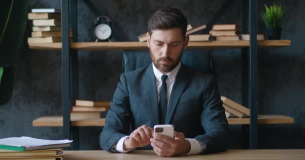 拥有现代智能手机的专业商人在办公室发短信。年轻男性雇员使用现代移动电话技术商业应用程序，坐在办公桌前输入电子邮件. — 图库视频影像