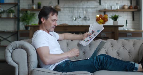 Side view of serious mature business man working with documents and laptop computer while lying on sofa at living room. Teleworking from home concept — Stock Video