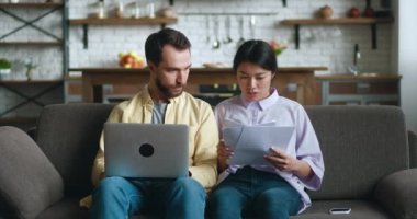 Çok ırklı genç bir çift, oturma odasındaki kanepede otururken iş belgeleriyle konuşup dizüstü bilgisayar kullanıyor. Beyaz adam ve Asyalı kadın bilgisayarlara bakıyor, evden telgraf çekiyor..