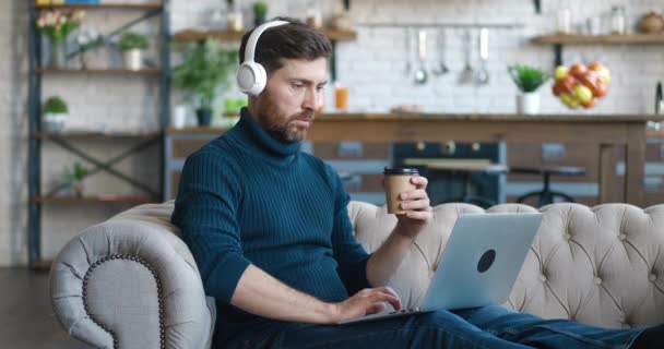 Schöner Millennial-Mann mit Kopfhörern, der am Laptop arbeitet und Kaffee trinkt, während er auf einem Sofa im gemütlichen Wohnzimmer sitzt. Freiberufler, die von zu Hause aus arbeiten. Surfen im Internet, Nutzung sozialer Netzwerke — Stockvideo