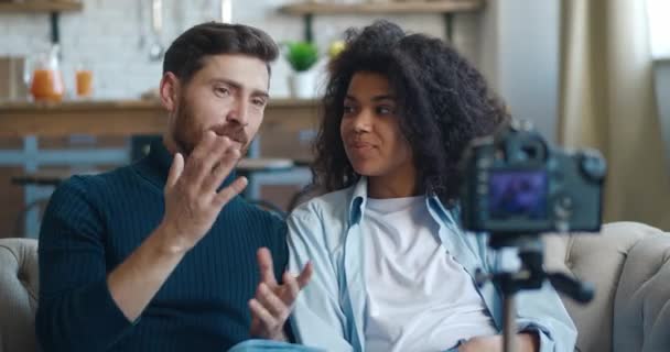 Jovens blogueiros familiares inter-raciais gravando vídeo para internet com câmera de vídeo profissional montada no tripé, sentados no sofá em casa. Homem e mulher conversando com câmera, gravando blog na sala de estar — Vídeo de Stock