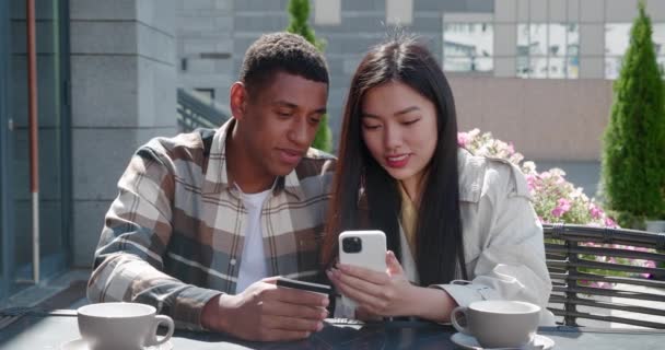 Jeune couple multi-ethnique de l'homme africain tenant une carte de crédit et femme asiatique gaie, assis à la table d'une terrasse d'été d'un café confortable, regardant le smartphone, faire des achats en ligne — Video
