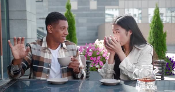 Charmantes multiethnisches verliebtes Paar sitzt an einem Tisch auf der Sommerterrasse eines gemütlichen Cafés, hält Tassen Tee in den Händen und unterhält sich bei einem Date an einem schönen warmen, sonnigen Tag gerne miteinander — Stockvideo