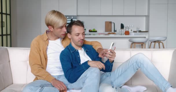 Ευτυχισμένοι άνδρες ομοφυλόφιλο ζευγάρι κάθονται στο σπίτι μαζί σε μια αγκαλιά στον καναπέ τους και χρησιμοποιώντας smartphone. Αρσενικό γκέι ζευγάρι περνάει χρόνο στο σπίτι. Σχέση ΛΟΑΤ — Αρχείο Βίντεο