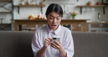 Akıllı telefon kullanan Asyalı genç bir kadın kazanan teklifi kutluyor, mutlu heyecanlı kız koltukta oturmuş akıllı telefona bakıyor iyi haberi kutluyor, başarı kazanan konsept