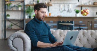 Yakışıklı Milenyum Adamı rahat oturma odasında bir koltukta otururken dizüstü bilgisayarda çalışıyor. Evden çalışan Freelancer. İnternet taranıyor, Sosyal ağlar kullanılıyor