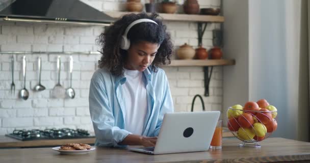 Freelancer feminino em fones de ouvido trabalhando em casa na cozinha e usando laptop. Uma jovem afro-americana a trabalhar com um computador. Menina estudante sentado e trabalhando no apartamento moderno. — Vídeo de Stock