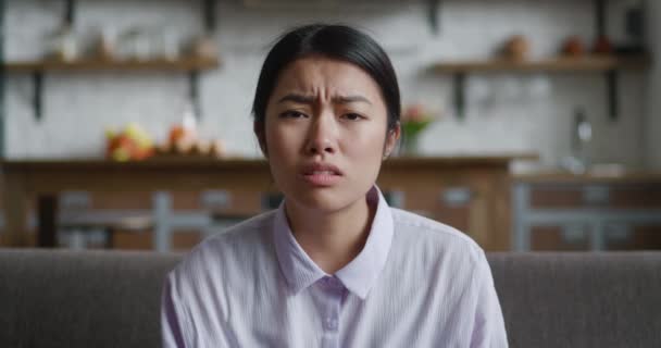 Verängstigte Asiatin schockiert. Attraktive junge Frauen schauen unangenehm überrascht in die Kamera, während sie zu Hause auf dem Sofa sitzen. Sie bedeckt ihren Mund mit einer Hand modernen Küchenhintergrunds — Stockvideo