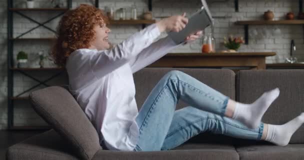 Вид сбоку на возбужденную рыжеволосую молодую женщину, сидящую на диване за ноутбуком, работающим в гостиной. Она получает отличные новости и в восторге. Показывать жест "да" — стоковое видео