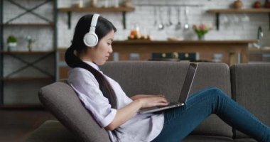 Kulaklıklı Asyalı bir kadın dizüstü bilgisayarla çalışıyor oturma odasındaki kanepede dinlenen ekran daktilo mesajlarına bakıyor, genç bir kadın internette sosyal medya okuyor ya da evde online çalışıyor.