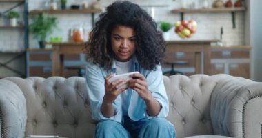 Komik Afrikalı Amerikalı kadın akıllı telefonuyla heyecan verici mobil uygulaması ile oynuyor. Gülümseyen kadın, evdeki oturma odasında kanepede otururken yumruğunu sıkarak bir yarış oyunu kazanıyor..