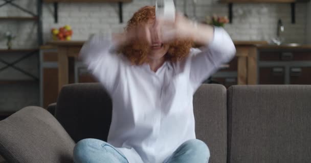 Mujer pelirroja joven y llena de alegría que usa el regalo ganador de un teléfono inteligente o el premio en la aplicación de redes sociales se sienta en el sofá en casa. Emocionado feliz mujer ganadora celebrando la victoria móvil en la sala de estar, mostrando sí gesto — Vídeo de stock