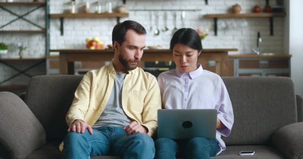 Счастливая многорасовая молодая пара разговаривает и использует ноутбук вместе смотреть видео, сидя на диване в гостиной. Кавказский мужчина и азиатская женщина смотрят на компьютер дома. — стоковое видео
