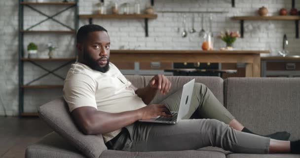 Афроамериканский бизнесмен на расстоянии работает на ноутбуке онлайн чата с сотрудниками компании на диване дома. Взрослый молодой человек фрилансер, лежащий на диване, использует компьютерную печать текста на клавиатуре — стоковое видео