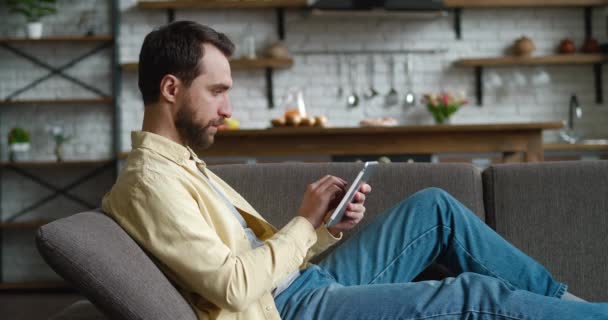 自宅でソファに休んでデジタルタブレットを使用して若い男,画面を見て若い男性がコンピュータを保持します。 — ストック動画