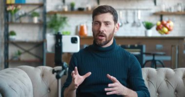 Blogcu adam evde akıllı telefon kamerasıyla video kaydediyor. Erkek kanepede oturuyor ve video konferansı veya video görüşmesi yapıyor. Vlog içeriği oluşturucu cep telefonuyla vlog kaydediyor