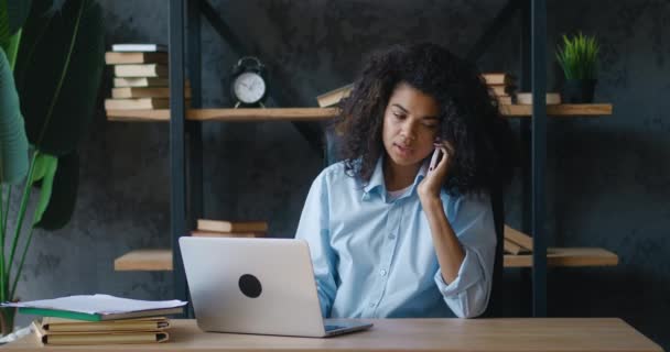 Afro-Amerikaanse zakenvrouw professioneel praten aan de telefoon terwijl ze aan het bureau zit. Jonge vrouwelijke student maken mobiel bellen tijdens het werken op laptop computer op moderne werkplek — Stockvideo