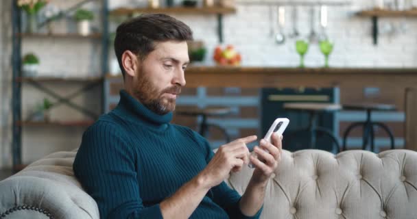 Vista lateral de hombre caucásico guapo sentado en el sofá en casa acogedora y el uso de teléfonos inteligentes navegar por Internet redes sociales, viendo videos y sonriendo. — Vídeos de Stock