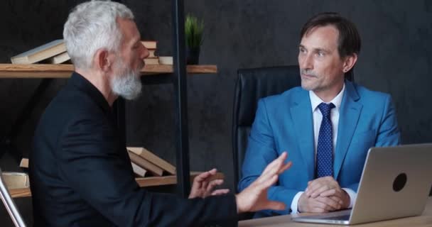 Two mature businessmen discussing business growth in modern office while sitting at desk. Man ceo talking with colleague about deal while meeting indoors — Vídeo de Stock