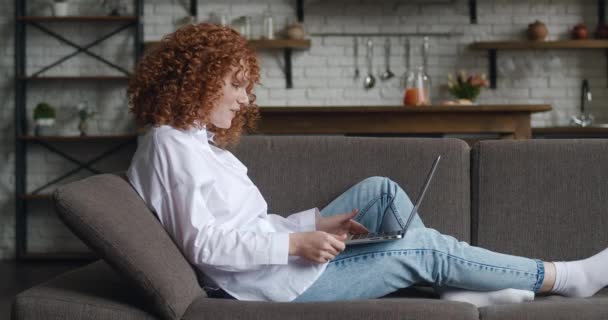 Vista laterale di felice donna dai capelli rossi con i capelli ricci che parla con il computer portatile, prendendo via chat virtuale online videochiamata mentre si siede sul divano a casa. — Video Stock