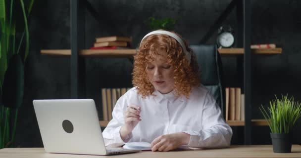 Kızıl saçlı, kulaklık eğitimi alan genç bir bayan öğrenci. İnternetteki öğretmen ile video konferansında sohbet ediyor. Video konferansını arıyor. — Stok video