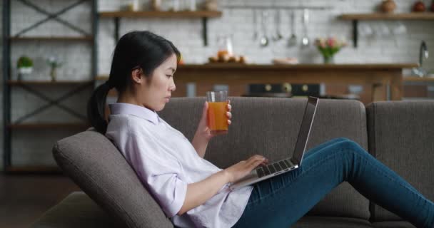 Mujer asiática bebiendo jugo descansando en el sofá usando el ordenador portátil mirando el mensaje de escritura de pantalla en la sala de estar, joven mujer charlando en la computadora navegando por Internet trabajando en línea en el sofá en casa — Vídeos de Stock
