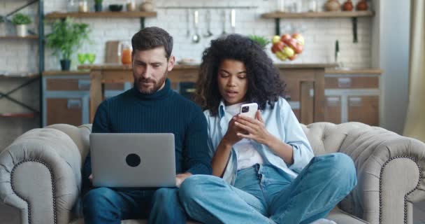 若い異人種間のカップルは、デジタルガジェットを使用して自宅でソファに休んでいます。スマートフォンを使用して彼のかなりアフリカ系アメリカ人の妻の間にノートパソコンの閲覧に取り組んで白人ハンサムな男。レジャーコンセプト — ストック動画