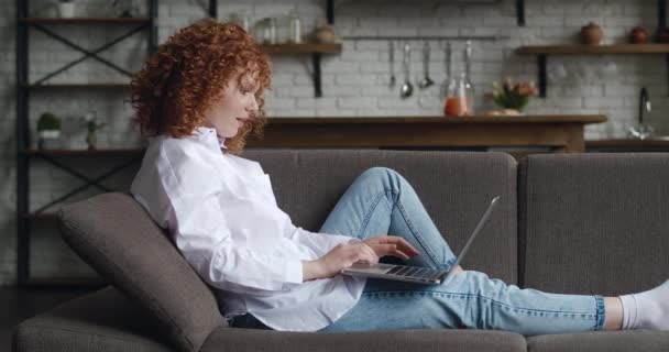 Vista lateral de la joven pelirroja feliz sentada en la acogedora sala de estar en el sofá con el uso de la computadora portátil. Sonriente estudiante chateando con amigos, trabajando o estudiando desde casa en línea en el sofá — Vídeo de stock