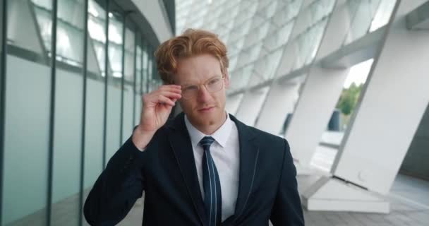 Der gut gelaunte junge rothaarige Geschäftsmann setzt seine Brille auf und blickt lächelnd in die Kamera, während er im Geschäftsviertel in der Nähe des Bürogebäudes steht. Im formellen Anzug. Porträt aus nächster Nähe. — Stockvideo