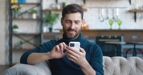 Bello uomo caucasico seduto sul divano a casa accogliente e utilizzando smartphone navigare su internet social media, guardare video e sorridere. — Video Stock