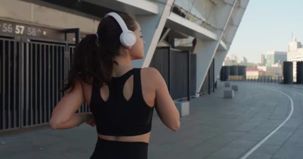 Widok z boku wysportowana młoda kobieta w słuchawki jogging i słuchanie muzyki na świeżym powietrzu w zwolnionym tempie. Fitness dziewczyna robi trening cardio, działa rano na ulicach miasta — Wideo stockowe