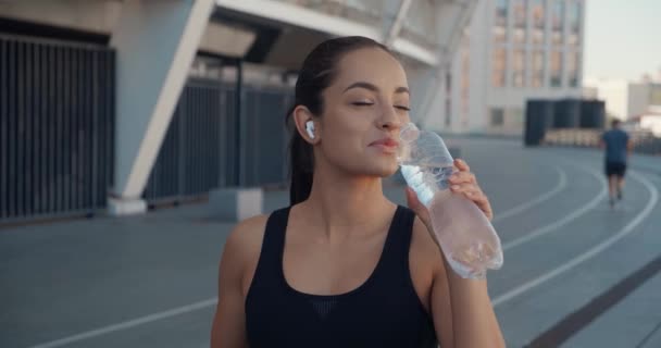 Piękna młoda wysportowana kobieta pijąca wodę z butelki po siłowni, trening na świeżym powietrzu, sport miejski. Brunetka fitness dziewczyna noszenie słuchawek, picie czystej wody mineralnej po joggingu na stadionie. — Wideo stockowe