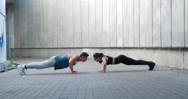 Pareja deportiva, hombre y mujer están haciendo flexiones juntos y tocándose las manos, al aire libre. Entrenamiento crossfit en grupo y concepto de estilo de vida saludable — Vídeos de Stock