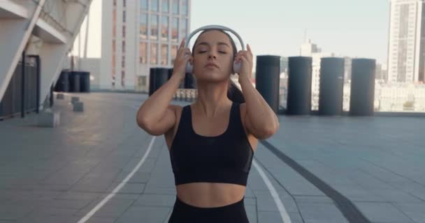 Widok z przodu wysportowana młoda kobieta umieścić w słuchawki i rozpocząć trening joggingu na świeżym powietrzu. Fitness dziewczyna robi trening cardio, bieganie rano na ulicach miasta i słuchanie muzyki — Wideo stockowe