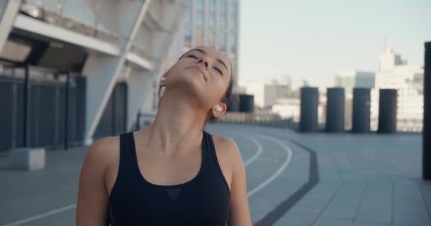 Retrato de bela jovem atlética usando fones de ouvido, relaxante após o treinamento esportivo, de pé ao ar livre. Morena menina fitness em sportswear olhando para a câmera no estádio. — Vídeo de Stock