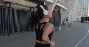 Kulaklıklı, koşan ve dışarıda ağır çekimde müzik dinleyen atletik bir genç kadın. Fitness kızı antrenman yapıyor, sabah şehir sokaklarında koşuyor.
