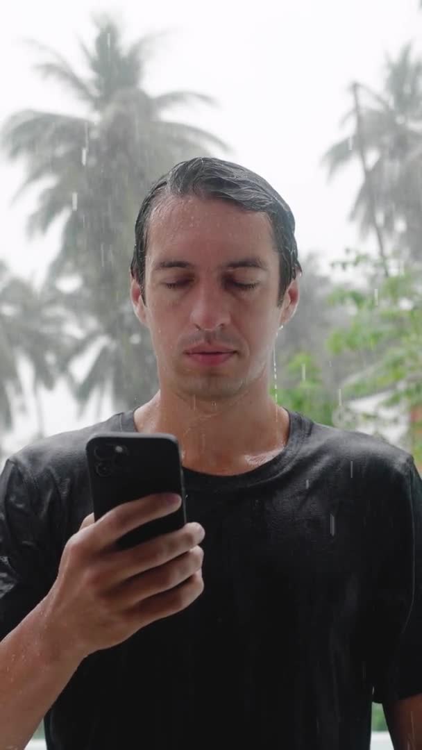 Weterynaryjny ujęcie mokrego młodego człowieka stojącego w ulewnym tropikalnym deszczu za pomocą swojego smartfona. Zwolniony ruch — Wideo stockowe