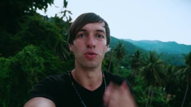 Yeşil orman arka planında selfie ya da video görüşmesi yapan gülümseyen genç bir turist. Genç erkek, görüntülü sohbet için tropikal bir ülkeyi geziyor..