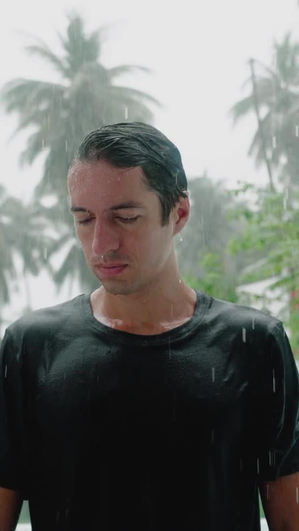 Retrato de un hombre pensativo y triste de pie bajo la lluvia y empapándose mientras mira a la cámara. En cámara lenta. Vídeo vertical — Vídeo de stock